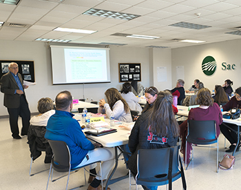At left, Vince Vandehaar, Principal Consultant at VVV Consulting, LLC, leads the group discussion at the Community Health Needs Assessment Town Hall Meeting.A group of several seated people listen as a presenter goes over local health statistics on a projected slide.